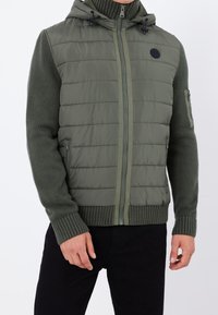 Veste à capuche rembourrée en vert olive avec un devant matelassé et des manches tricotées. Comprend une fermeture éclair à l'avant, des poches latérales et un patch logo.