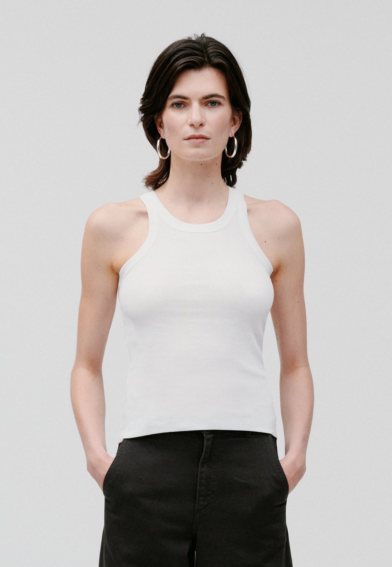Top din bumbac alb, cu design de tip racerback și siluetă ajustată, asociat cu pantaloni negri. Textură netedă, detalii minime.