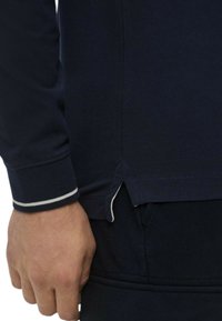 Camicia a maniche lunghe blu navy con polsini a righe bianche, spacchi laterali all'orlo e tessuto strutturato. Mano visibile che afferra il tessuto.