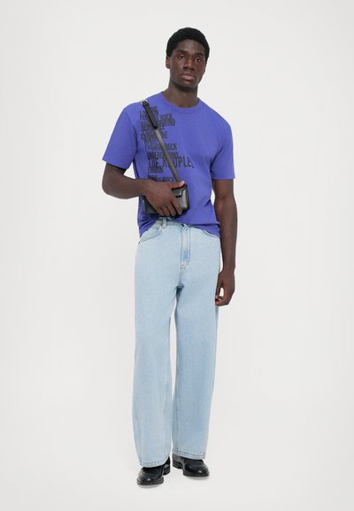 Purple t-shirt with black text, light blue wide-leg jeans, and black boots. Model holds a small black bag. Simple, minimalistic design.
