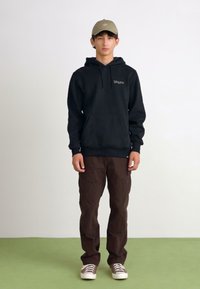 Black hooded sweatshirt with a front pocket, paired with brown cargo pants and beige sneakers. The cap is light green with a logo.