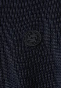 Bonnet en tricot bleu marine foncé avec un motif texturé. Présente un écusson rond noir avec un design géométrique.