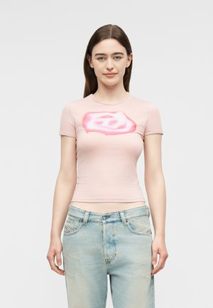 Νεαρή γυναίκα με μακριά καστανά μαλλιά φοράει ένα ανοιχτό ροζ εφαρμοστό t-shirt με κόκκινο αφηρημένο σχέδιο και ανοιχτό μπλε τζιν παντελόνι.