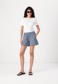 Femme portant des lunettes de soleil, un haut blanc à manches courtes, un short à carreaux bleu et blanc, et des tongs noires, se tenant devant un fond blanc uni.