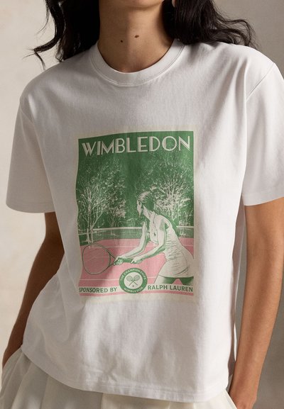 Γυναίκα που φοράει λευκό T-shirt με πράσινο και ροζ γραφικό μιας θηλυκής τενίστριας που κρατά ρακέτα στο γήπεδο, με το κείμενο Wimbledon από πάνω.