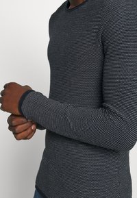Chemise tricot à manches longues, bleu marine, avec un motif à pois blancs, au design ajusté, col rond et poignets noirs contrastants. Tissu texturé.