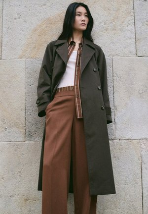 Mujer con abrigo largo verde oscuro sobre camisa marrón a rayas, camiseta blanca y pantalones marrones de pierna ancha, de pie junto a una pared de piedra.