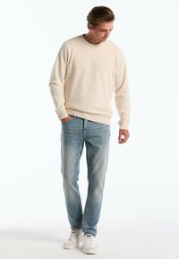 Crème-kleurige sweatshirt met een ronde hals, geribbelde boorden, gecombineerd met lichtblauwe spijkerbroek. Model draagt witte sneakers. Eenvoudig ontwerp.