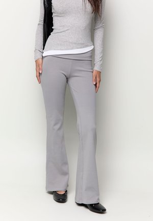 Femme portant un pantalon évasé gris clair, un haut gris à manches longues, des chaussures noires et portant un sac à épaule noir, debout devant un fond blanc.