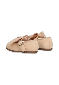 Zapatos de bebé en color beige con lazos de satén, correas ajustables con hebillas laterales y suelas planas marrones, mostrados desde un ángulo trasero sobre un fondo blanco.