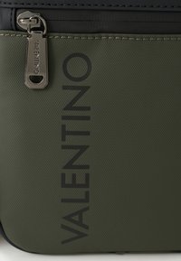 Πράσινη υφασμάτινη τσάντα με υφή, με κλείσιμο με φερμουάρ, εμφανές λογότυπο 'VALENTINO' σε μαύρο χρώμα και ασημένια μεταλλικά εξαρτήματα.