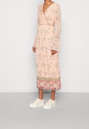Femme portant une robe portefeuille à manches longues, rose pâle avec des motifs floraux et un ourlet à motifs, associée à des baskets blanches à plateforme.