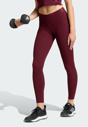 Kvinde i vinrøde leggings og crop top, der holder en sort Adidas håndvægt, iført sorte og hvide Adidas sneakers, stående på en lys baggrund.