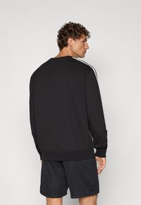 Sudadera negra de manga larga, con cuello redondo y rayas blancas en los laterales. La tela parece suave; tiene un corte relajado, combinada con pantalones cortos oscuros.