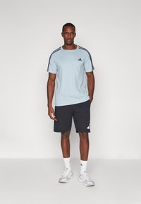 Világoskék póló fekete Adidas logóval és vállrészen fekete csíkokkal, sötétszürke rövidnadrággal párosítva, amelyen fehér Adidas akcentus található.