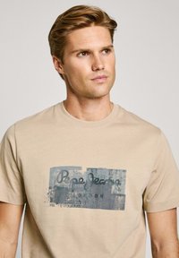 Bežinė medvilninė marškinė su mėlynu grafikiniu dizainu, kuriame užrašyta "Pepe Jeans London". Marškiniai su apvalia iškirpte ir trumpomis rankovėmis.