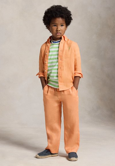 Niño con cabello rizado está de pie con las manos en los bolsillos, lleva una camisa y pantalones naranjas, una camisa de rayas verdes y blancas, y zapatos oscuros sin cordones.