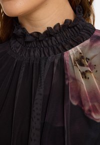 Zwarte mesh-blouse met rimpelige hoge hals, voorzien van doorschijnende stof en bloemmotiefdetails in gedempte kleuren. Subtiele textuur en gelaagd ontwerp.