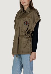 Gilet verde oliva, in tessuto leggero, design oversize con bordi sfrangiati, multiple tasche, vita regolabile con coulisse, con una toppa circolare sul petto.