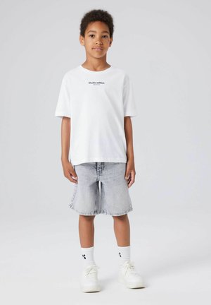 Garçon debout, portant un T-shirt blanc "Studio edition", un short en jean gris clair, des chaussettes blanches et des baskets blanches sur fond gris uni.