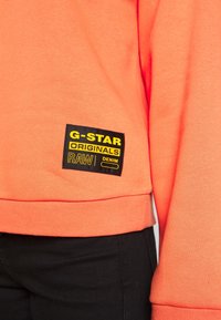 Korálová mikina s žebrovaným lemem a manžetami. Na ní je černá obdélníková štítek se žlutým nápisem: "G-STAR ORIGINALS RAW DENIM."