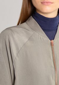 Femme portant une veste beige zippée sur un col roulé bleu marine, montrant le haut de la poitrine et les lèvres, avec des cheveux bruns raides visibles du côté gauche.