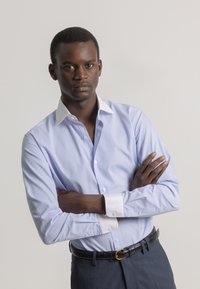 Camisa de botones azul claro con cuello y puños blancos, de tejido texturizado, combinada con pantalones oscuros y un cinturón negro con una hebilla dorada.