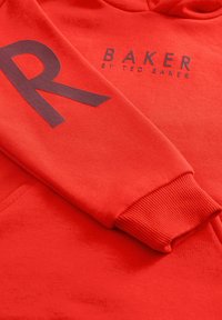 Sarkana kapuce ar lielu melnu "R" uz piedurknes un tekstu "BAKER by TED BAKER". Izgatavota no teksturēta auduma ar ribotām piedurknēm.