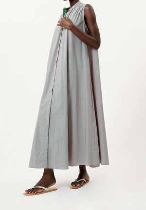 Nainen hihattomassa, nilkkaa myöten ulottuvassa raidallisessa mekossa, jossa on napit, pitäen kiinni kauluksesta, pukeutuneena beigen värisiin tasapohjaisiin sandaaleihin, seisomassa valkoista taustaa vasten.