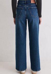 Vue arrière d'une personne portant un jean bleu taille haute et large, accompagné d'un haut noir à manches longues, sur un fond uni.