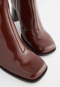 Bottes de cheville en cuir verni marron avec un bout carré, un talon modéré et des détails de couture visibles le long de la couture. Fini lisse et brillant.