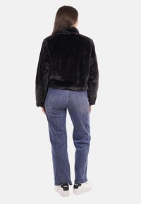 Giacca in pelliccia sintetica nera con colletto alto, abbinata a jeans blu e sneakers bianche. La giacca presenta una finitura morbida e testurizzata.