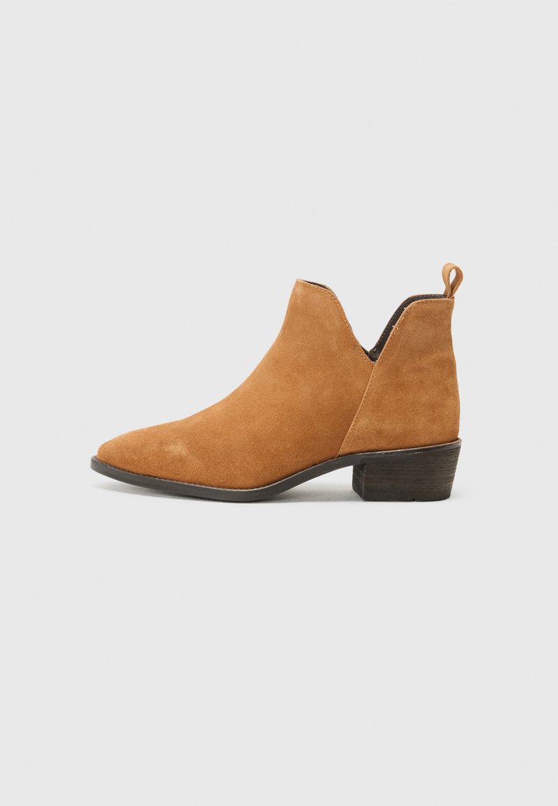 Bottines en suede beige à bout pointu, avec un petit talon empilé et un design découpé sur les côtés. Elles possèdent une petite boucle arrière pour un enfilage facile.