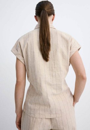 Mujer con coleta larga marrón que lleva una camiseta beige de manga corta y pantalones a juego con finas rayas verticales, de pie con una mano en la cadera.