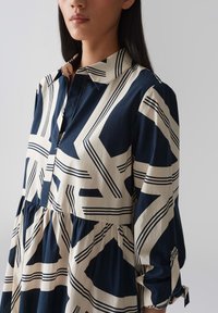 Navy and cream patterned button-up dress, featuring geometric designs, a collar, and gathered waist with long sleeves and cuff details.