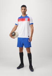 Hvid Nike fodboldshirt med blå krave og ærmer, med en rød gradient og Englands våbenskjold; parret med blå shorts og sorte sokker.