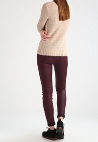 Beige Strickpullover mit geripptem Saum, kombiniert mit eng anliegenden bordeauxroten Hosen. Schwarze Stiefeletten mit braunen Akzenten, umgeschlagene Hosenenden.