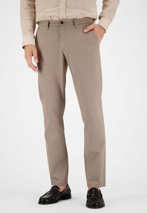 Mann trägt beige strukturierte Hose, hellbeiges Hemd mit Knöpfen und hochgekrempelten Ärmeln sowie schwarze Leder-Loafer, steht mit einer Hand in der Tasche.