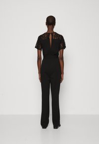 Combinaison noire avec des manches courtes en dentelle, un détail de dos en trou de serrure et un pantalon large, fabriquée en tissu lisse avec une taille cintrée.
