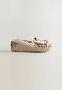 Chaussure d'intérieur beige en matériau doux, avec un accent en nœud, un dessus texturé avec de petits points, et un bord cousu le long de la semelle.