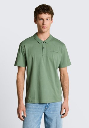 Man draagt een groen poloshirt met de tekst "Denim Design Studio" en lichtblauwe spijkerbroek, staand tegen een effen grijze achtergrond.