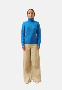 Maglione a collo alto blu lavorato a maglia con una texture a coste, abbinato a pantaloni larghi in velluto a coste beige con strisce verticali.