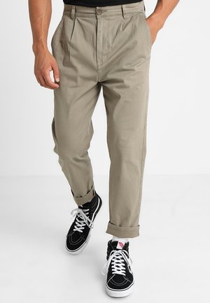 Uomo che indossa pantaloni beige risvoltati e sneaker nere alte con lacci bianchi, che cammina con le mani in tasca.