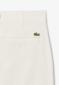 Chino in cotone beige con una tasca posteriore con chiusura a bottone bianco e un logo coccodrillo ricamato verde sul lato destro.