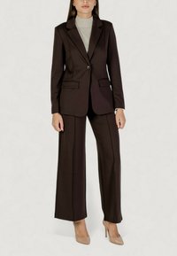 Traje marrón a medida con blazer de un solo botón, solapas con muesca y dos bolsillos frontales, combinado con pantalones de pierna ancha y tacones nude.