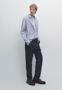 Light blue striped long-sleeve shirt, black belt, dark denim pants, and brown shoes. Model is standing against a plain background.