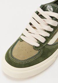 Grønn og brungul suede sneakers med hvite snører og sømmer, med et klassisk design og en teksturert gummisåle.