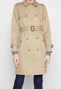 Beige trenchcoat med dubbelknäppta knappar, bälte vid midjan, sidofickor och manschettremmar. Tillverkad av ett texturerat tyg.