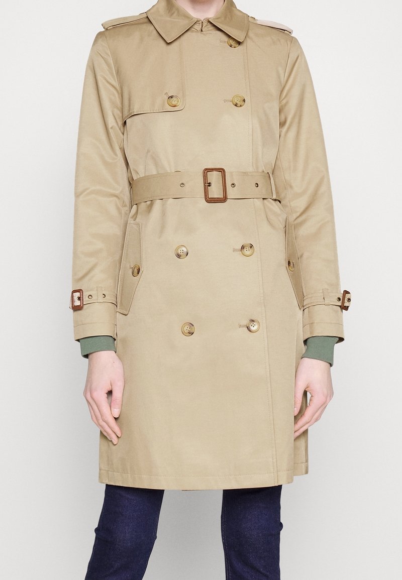 Beige trenchcoat med dubbelknäppta knappar, bälte vid midjan, sidofickor och manschettremmar. Tillverkad av ett texturerat tyg.