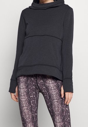 Mujer con sudadera con capucha de manga larga en gris oscuro y leggings de cintura alta con un patrón de piel de serpiente en púrpura y negro.